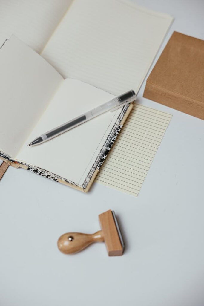 A minimalist workspace featuring an open notebook, pen, and wooden stamp on a white surface.